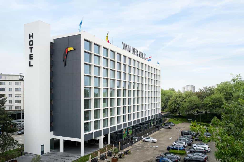 Modern Van der Valk Hotel building exterior with parking lot and surrounding green trees.