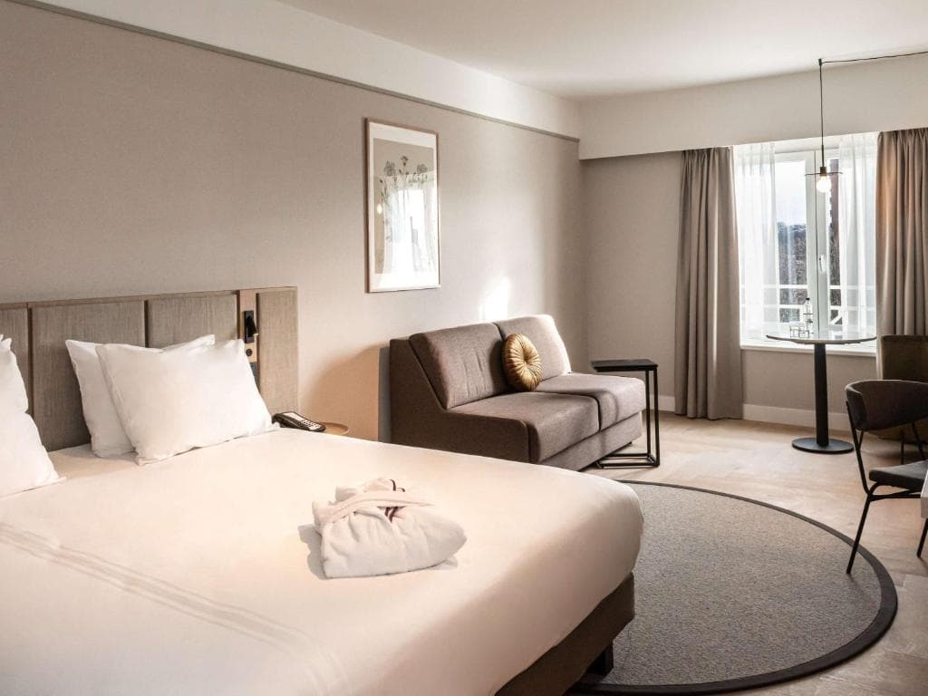 Modern hotel room with large bed, sofa, and small dining table near a window