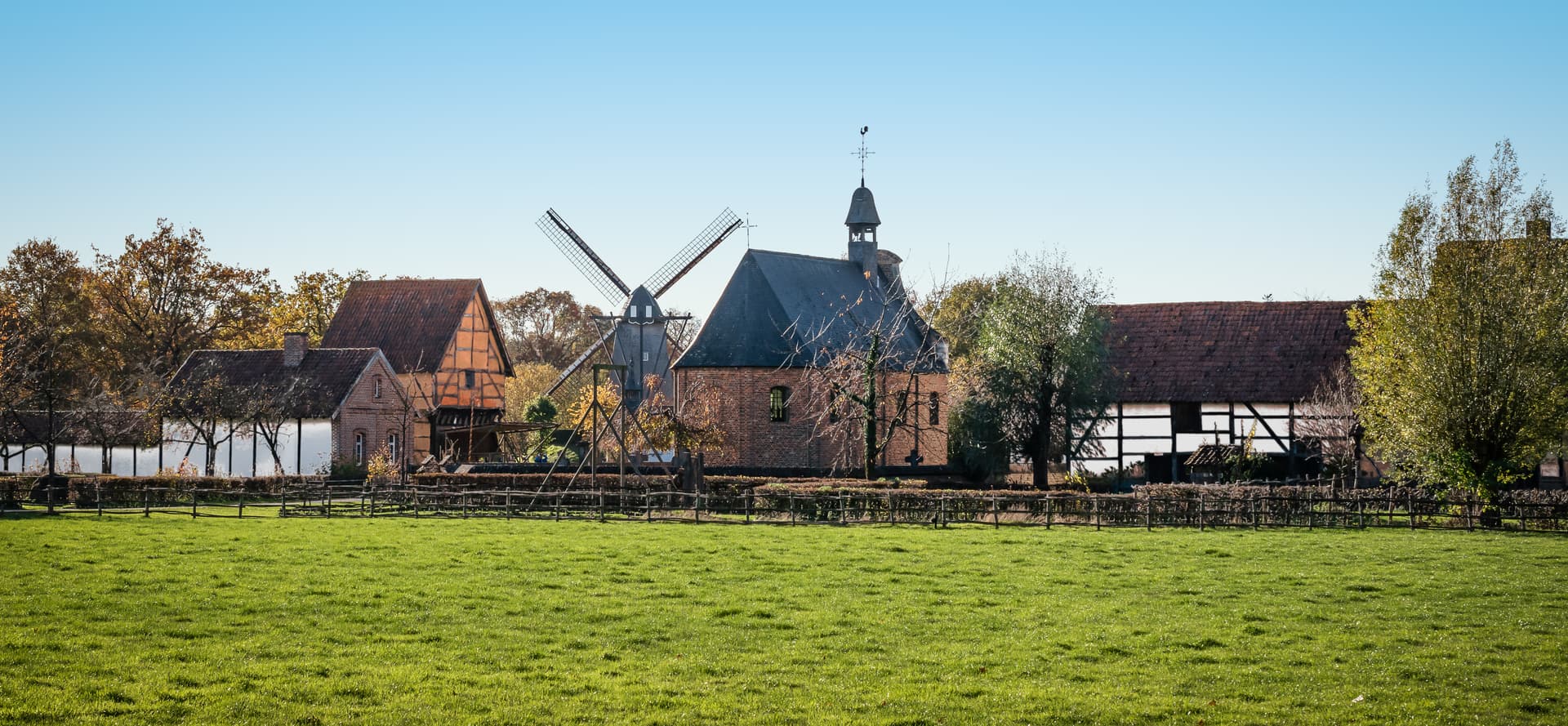 Windmill and historic buildings behind a bright green field under a clear blue sky.