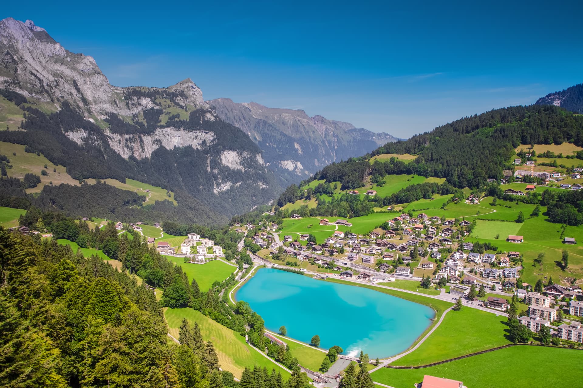 Alpine village nestled by bright turquoise lake with steep green mountains under clear blue sky.