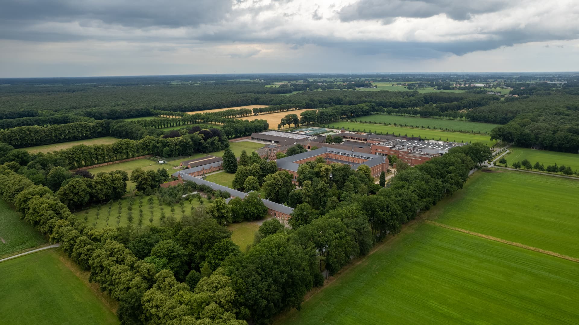 Aerial view of Westmalle Abbey complex surrounded by green fields and dense forest under cloudy skies.