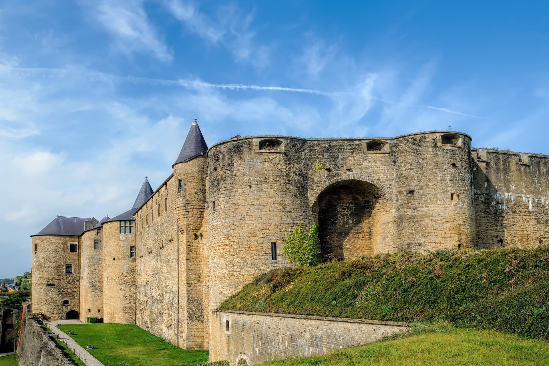 Stone fortress walls with conical towers and green grassy ramparts under a blue sky.