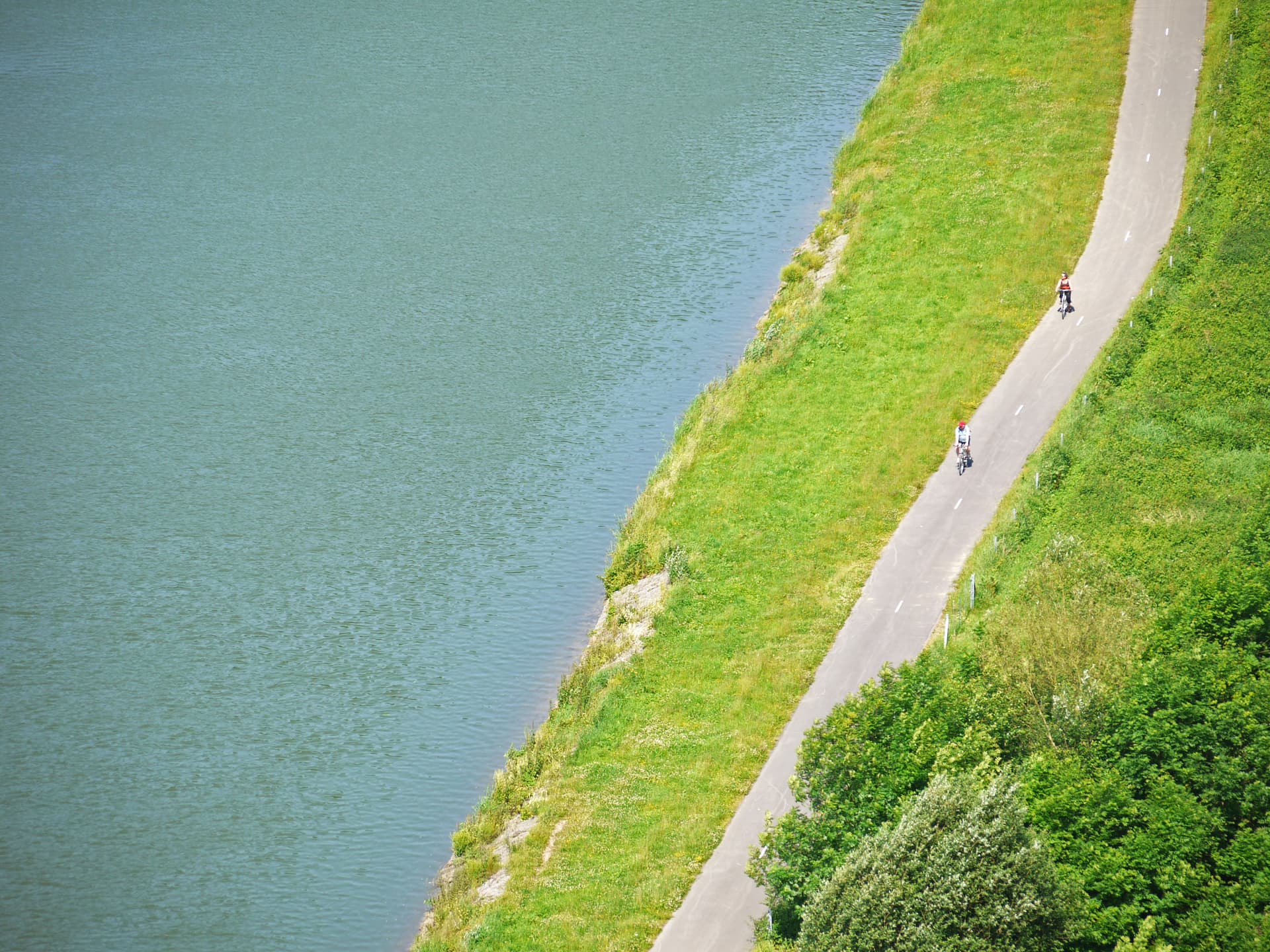 Cyclists ride paved path alongside green bank and calm river water, Meuse route.