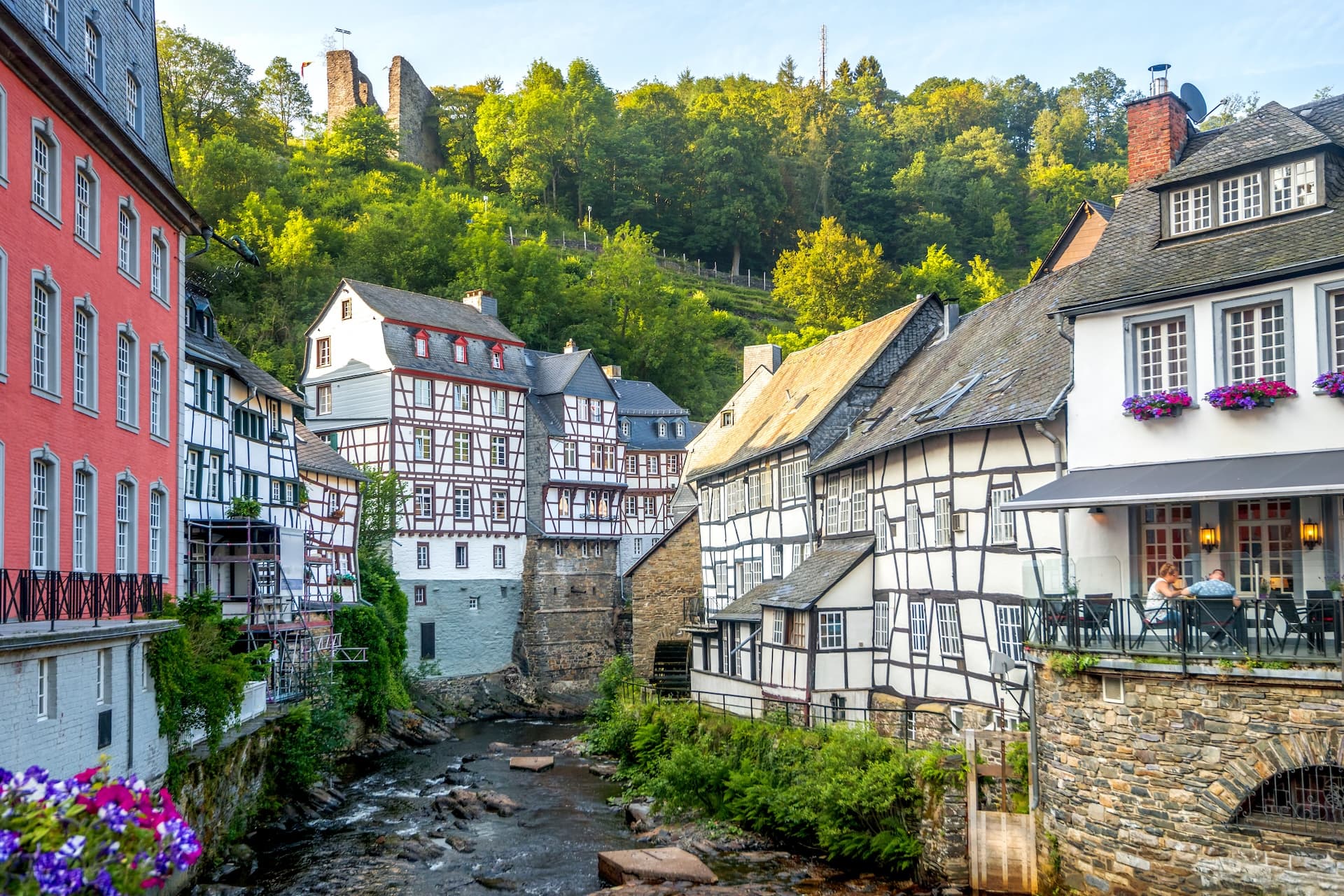 Half-timbered houses along a rocky stream with ruins on a forested hill in Monschau.