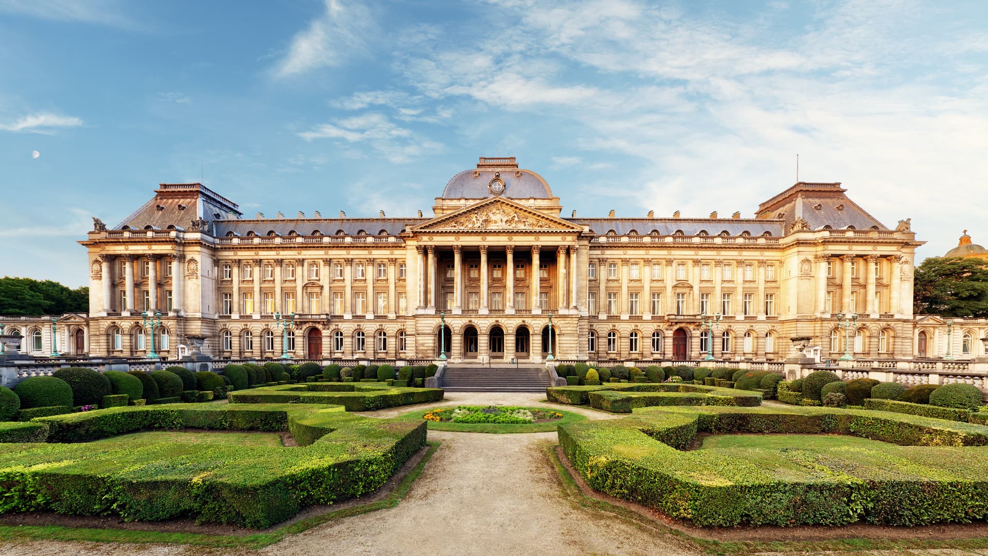 Royal Palace of Brussels facade with formal gardens and manicured hedges under a blue sky.