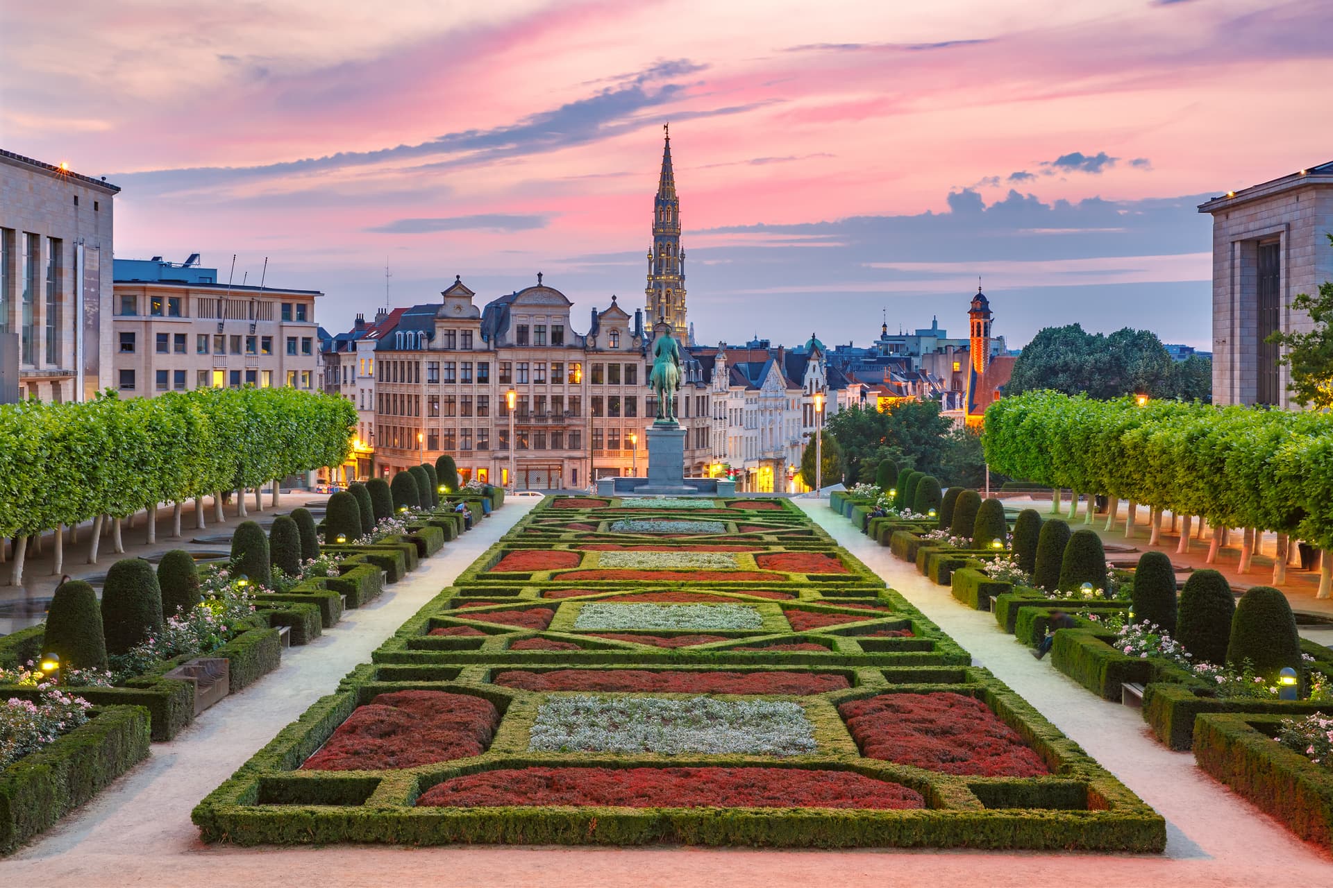 Formal garden with patterned hedges at dusk, overlooking city buildings and a spire.
