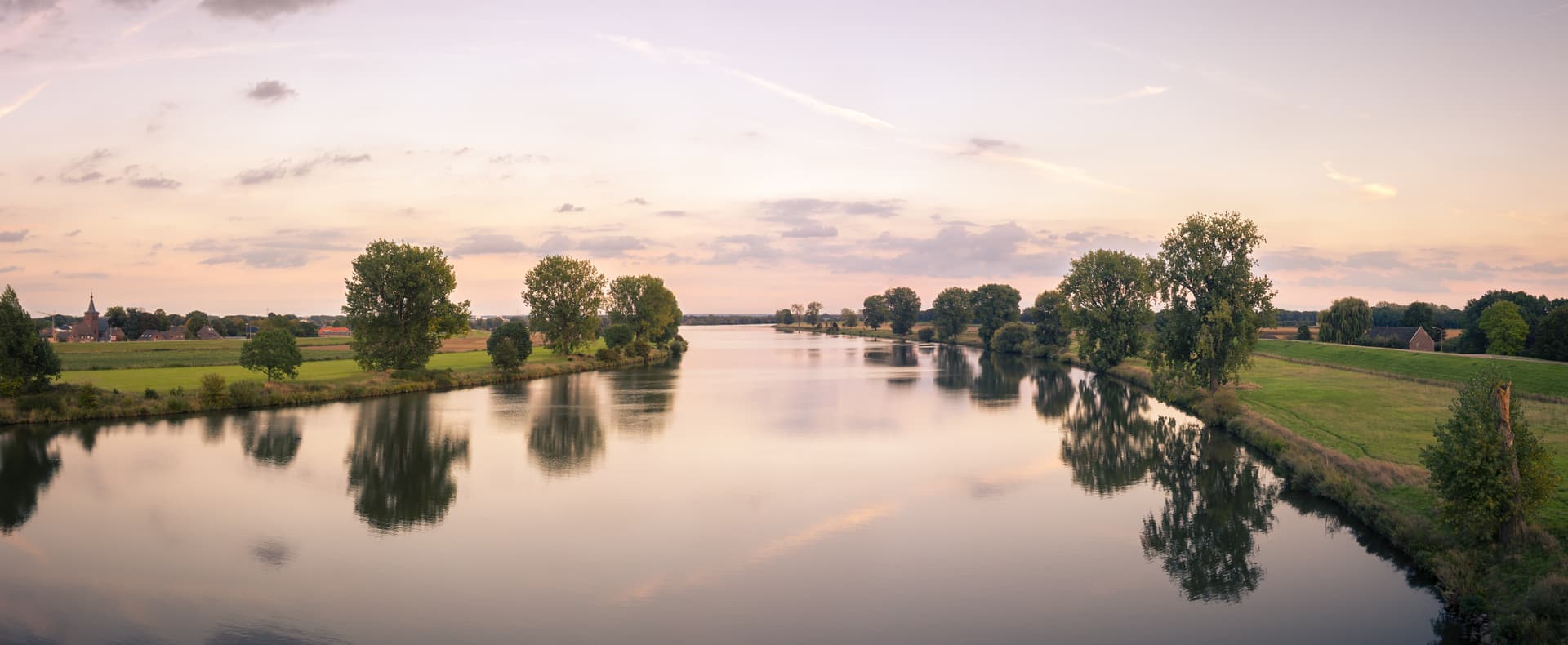 Calm river reflecting trees and pastel sunset sky near rural village buildings.