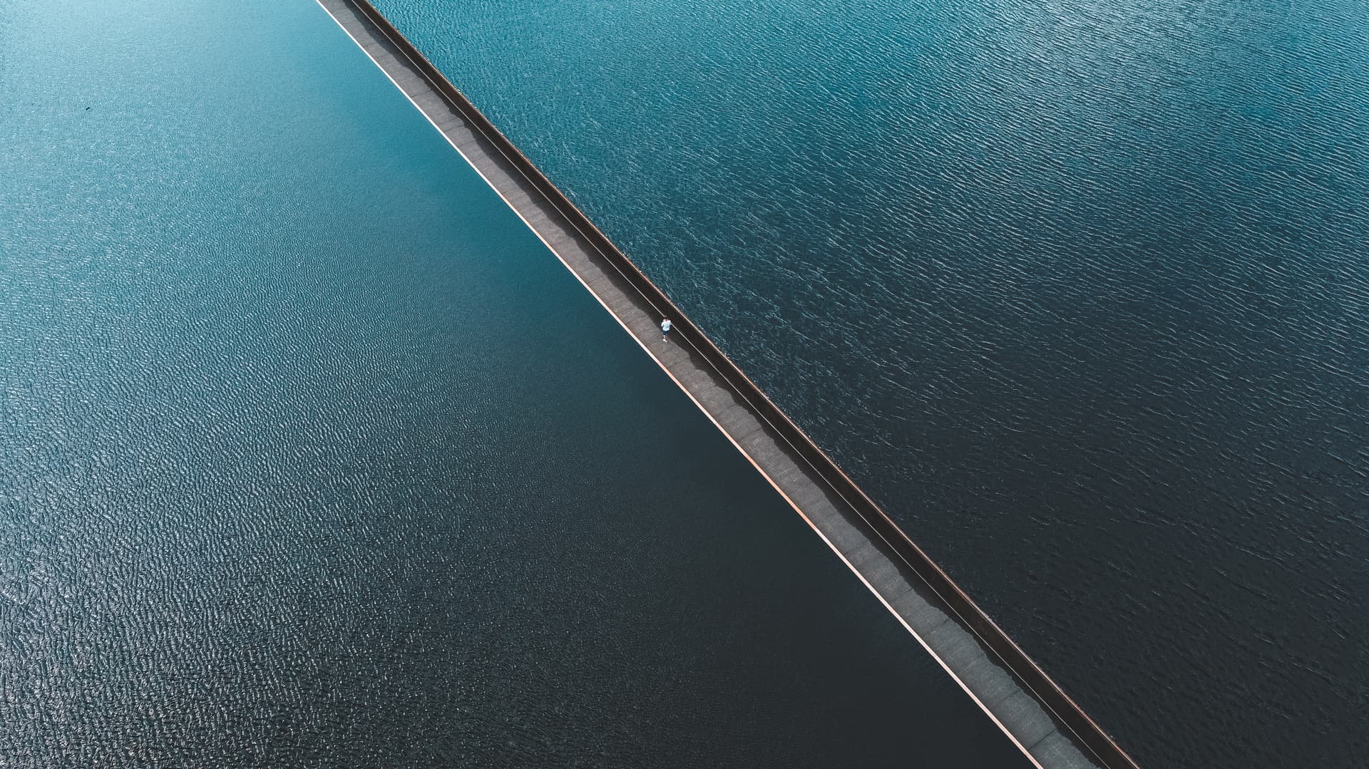 Person walking on narrow walkway separating two bodies of dark blue water from above.
