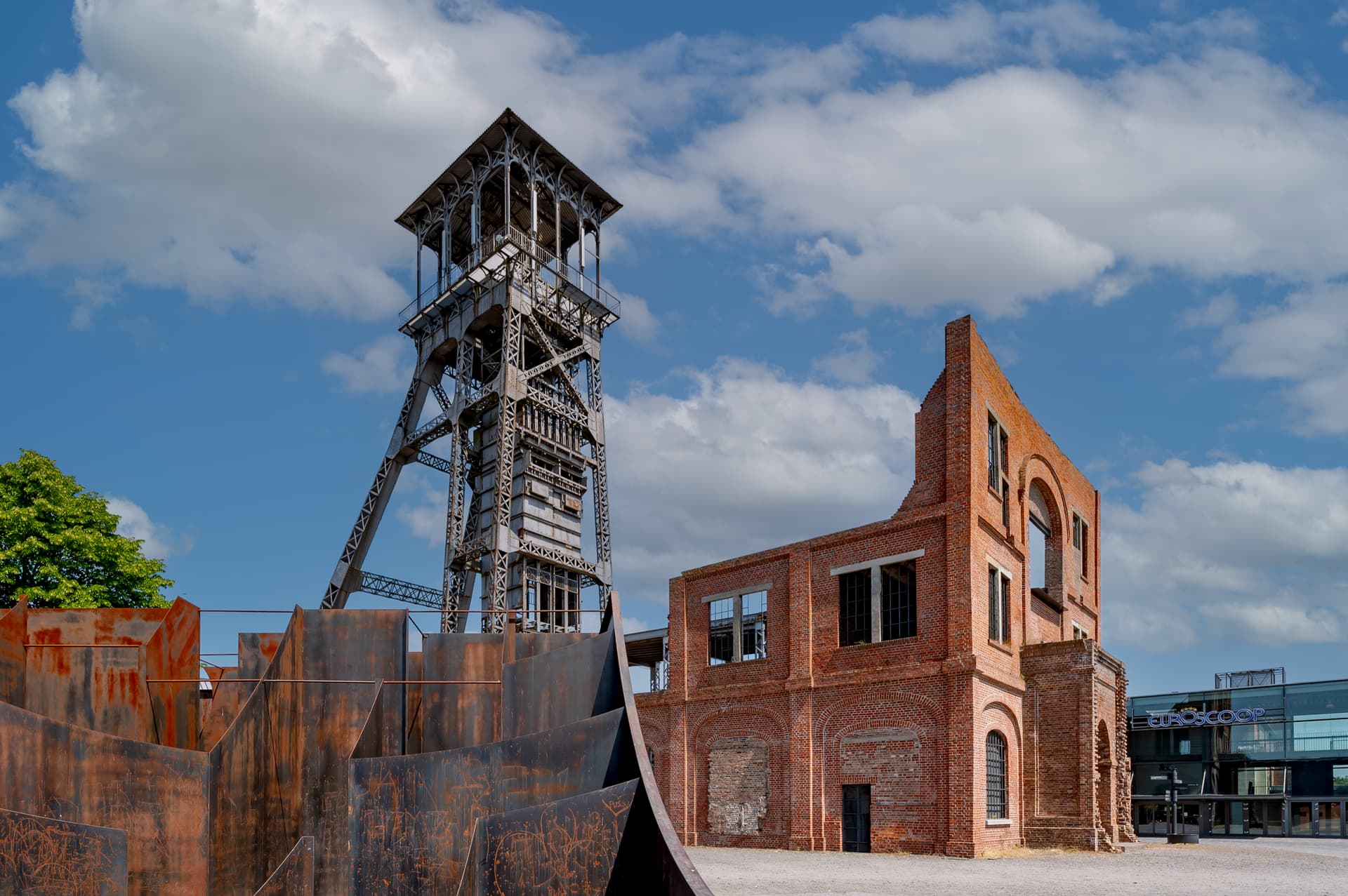 Industrial headframe tower next to ruined brick building and rusted metal sculpture under blue sky.