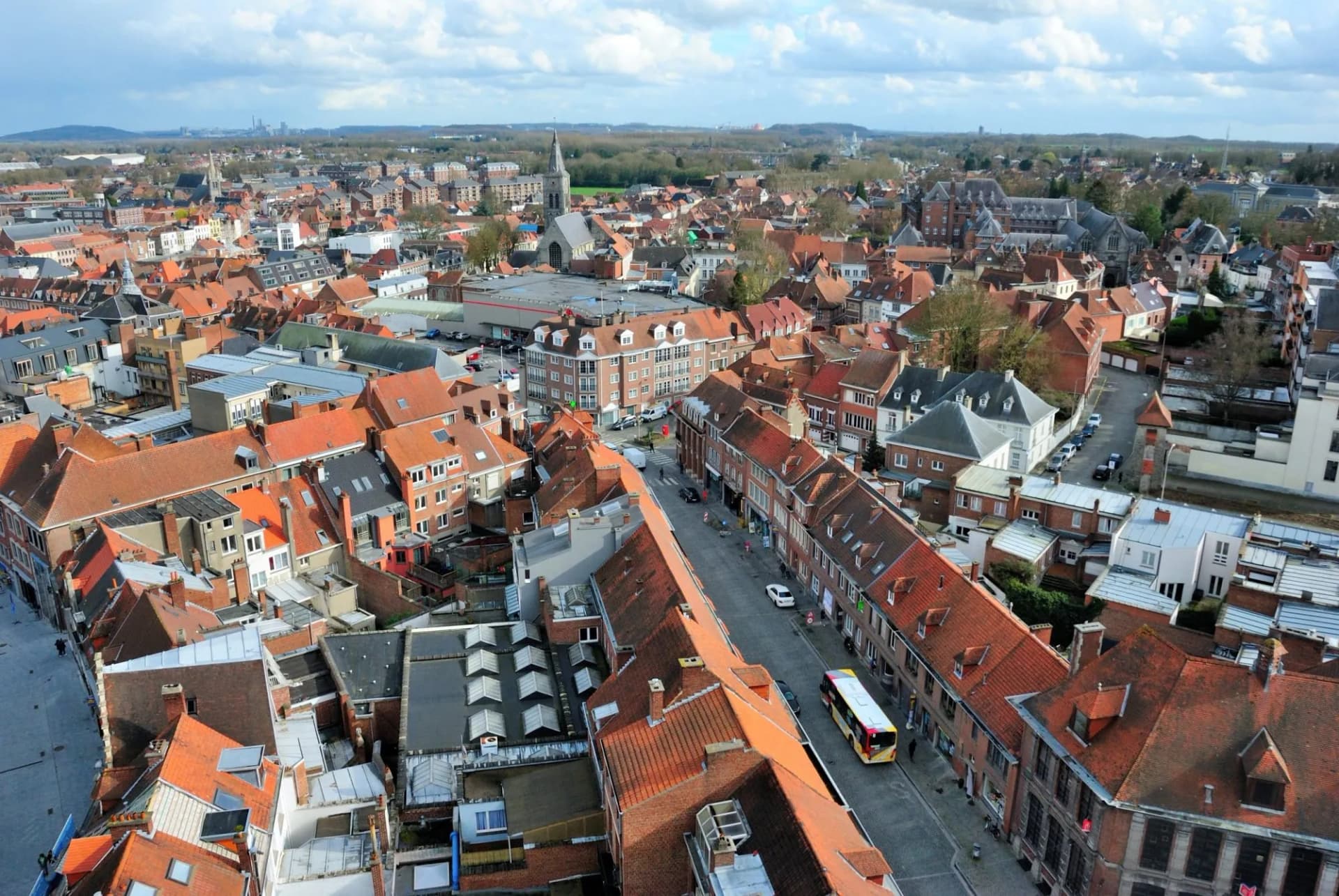 Aerial view of dense European town with red tile roofs, a church spire, and a bus on the street.