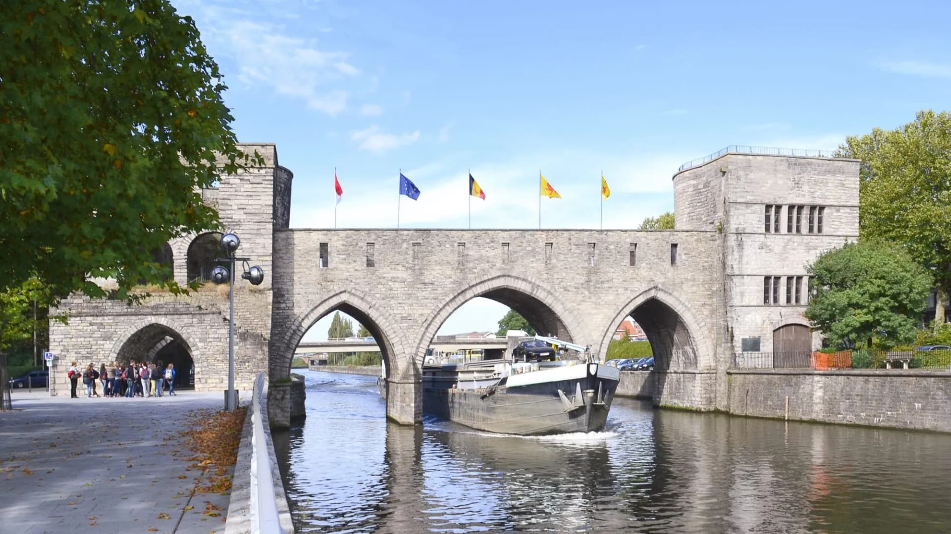Cargo boat passes under historic stone bridge with flags flying above river in Doornik.