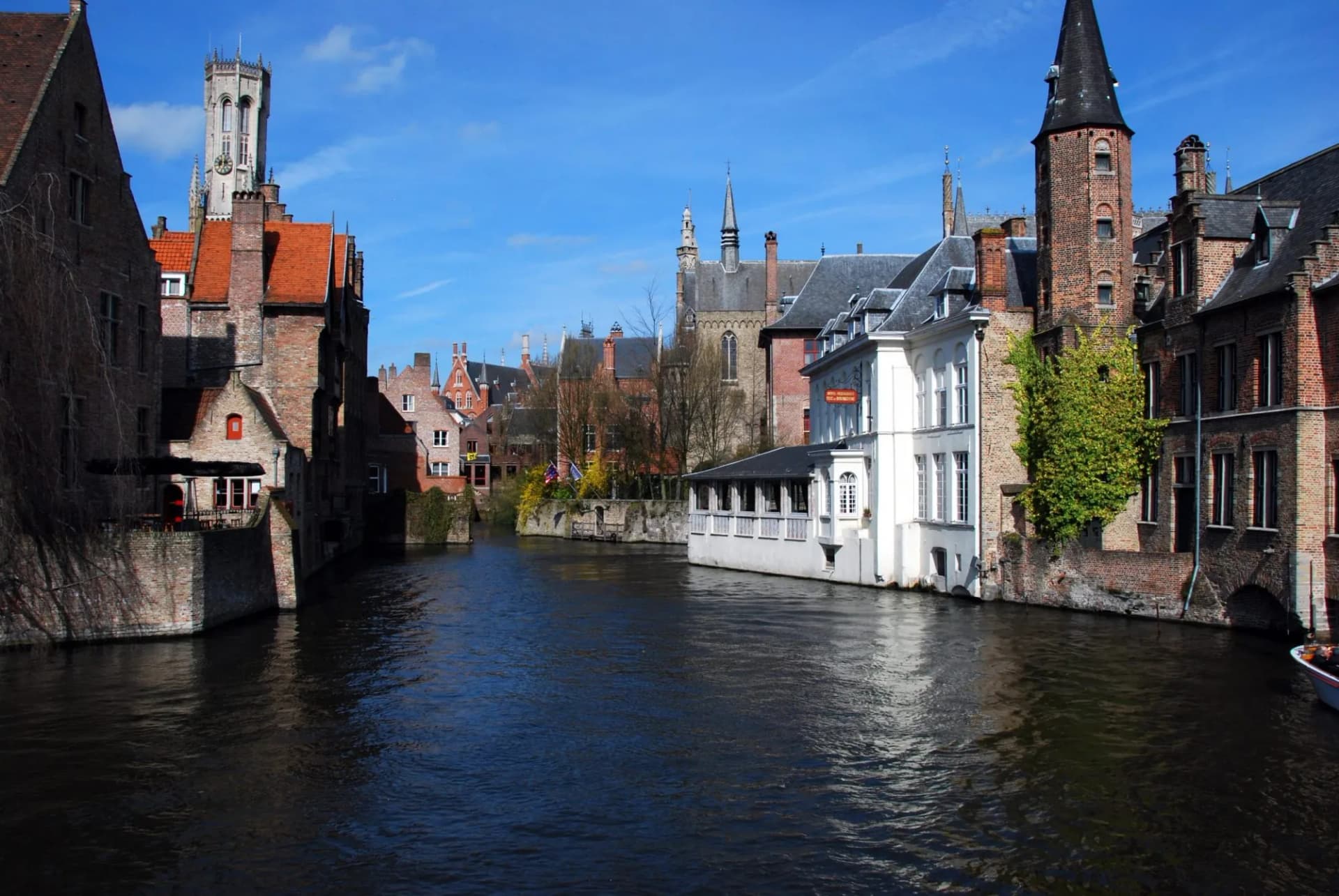 Canal with historic brick buildings and Belfry tower under blue sky in Bruges.