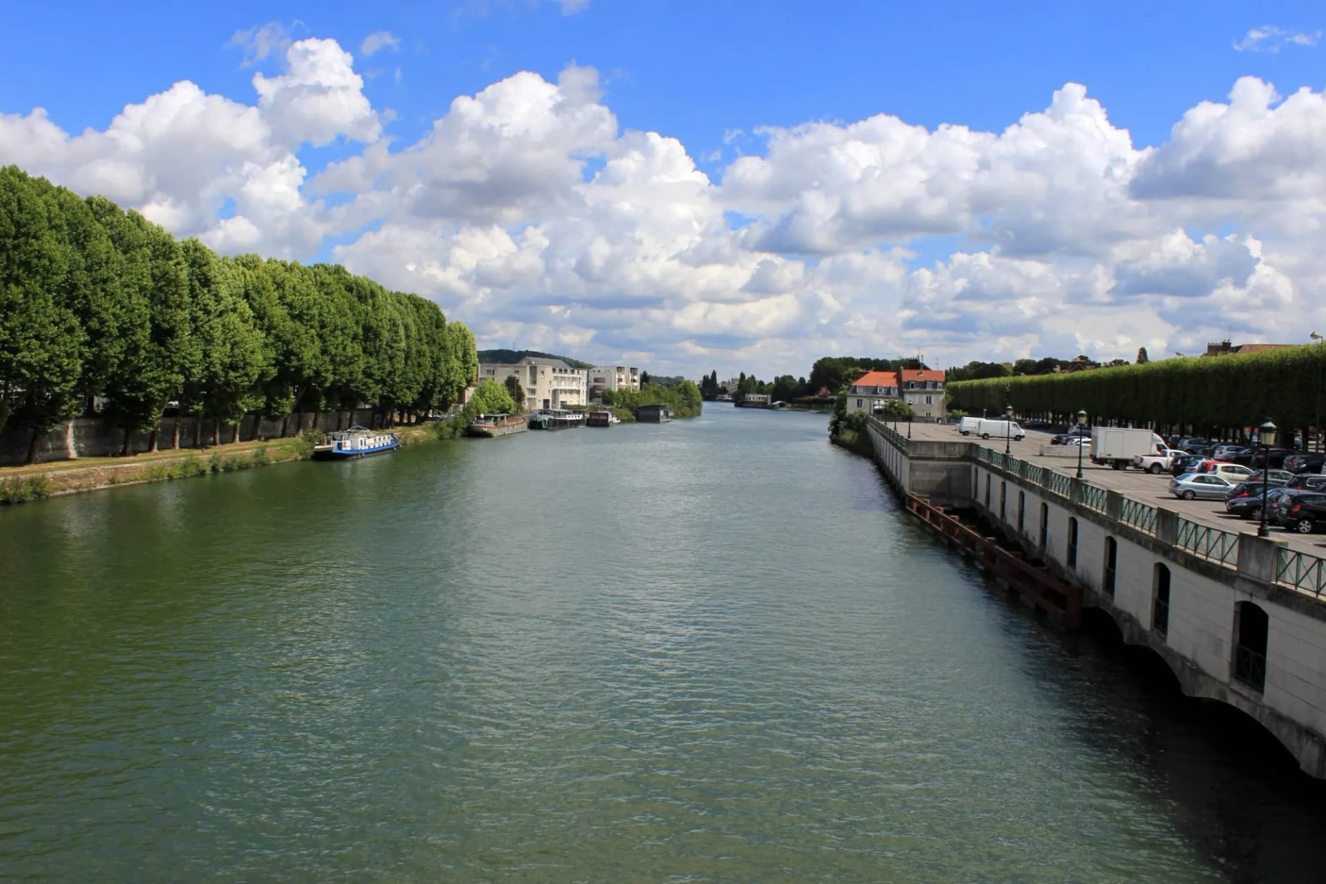 River in Compiègne with tree-lined banks and a docked barge under a blue, cloudy sky.