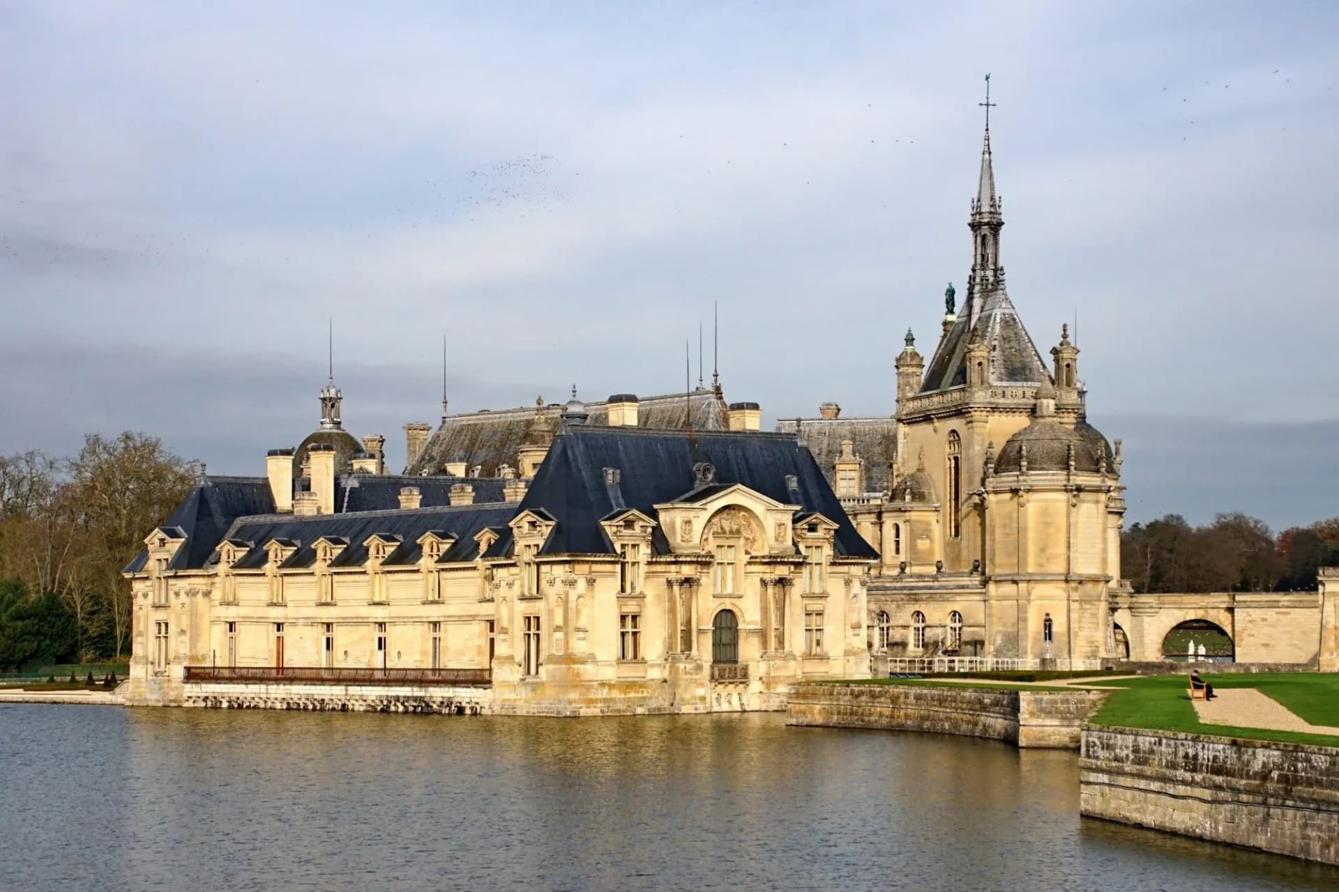 Château de Chantilly reflected in the water with formal gardens and a person sitting on a bench.