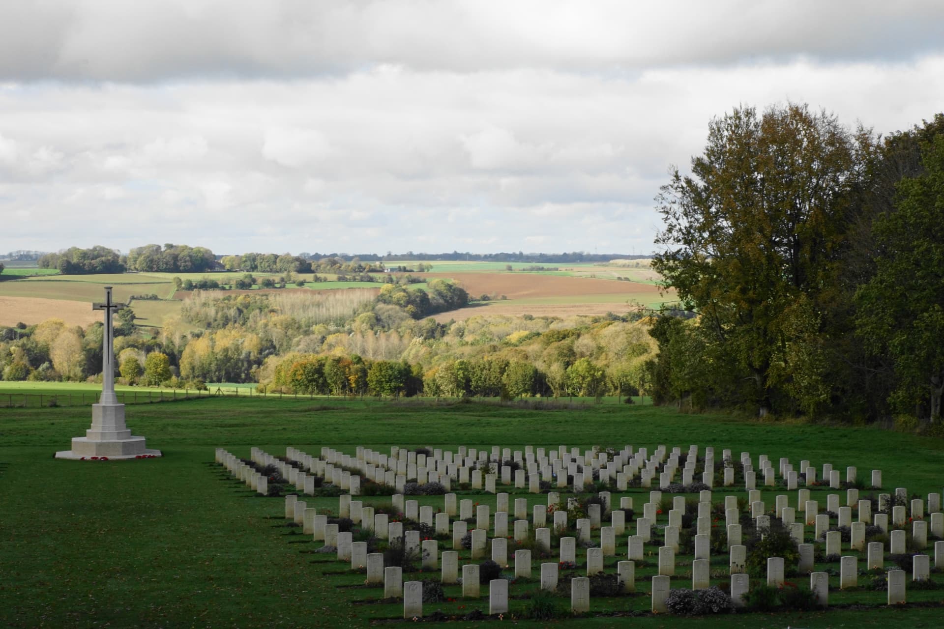 War cemetery with rows of white headstones and a large stone cross overlooking rolling fields.