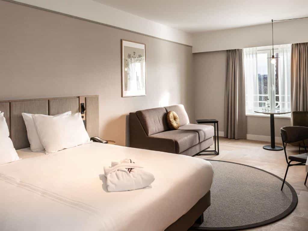 Modern hotel room with large bed, sofa, and small dining table near a window