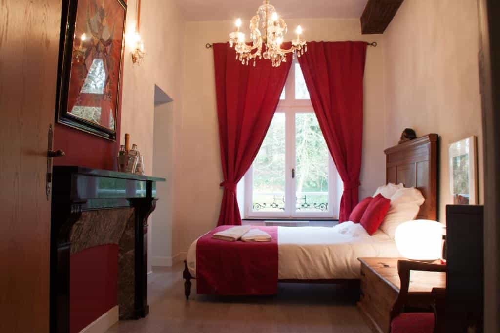 Bedroom with wooden bed, red accents, crystal chandelier, and fireplace, looking out a window.
