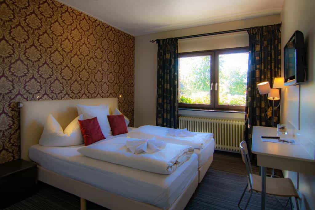 Twin beds with white linens, red pillows, and damask wallpaper in a hotel room.