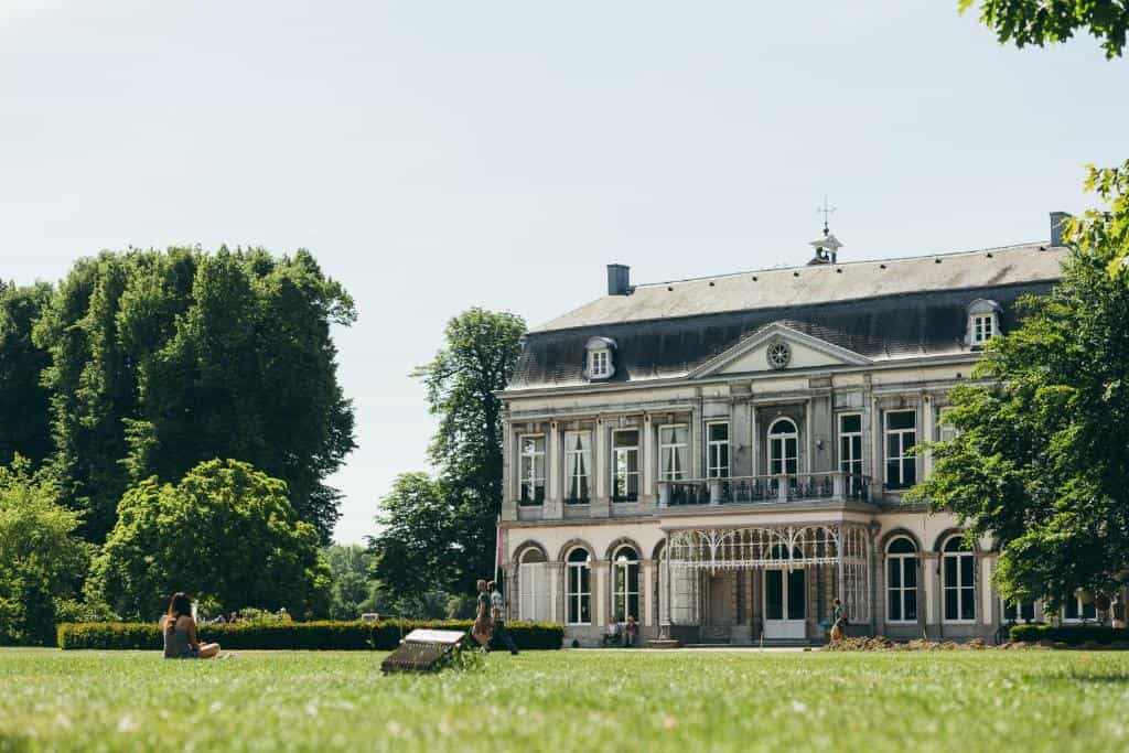 Stone chateau with large windows and dark roof surrounded by lush green trees and lawn.