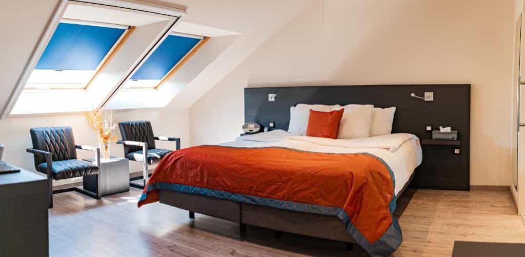 Attic bedroom with sloped ceiling, skylights, large bed with orange duvet, and two dark armchairs.