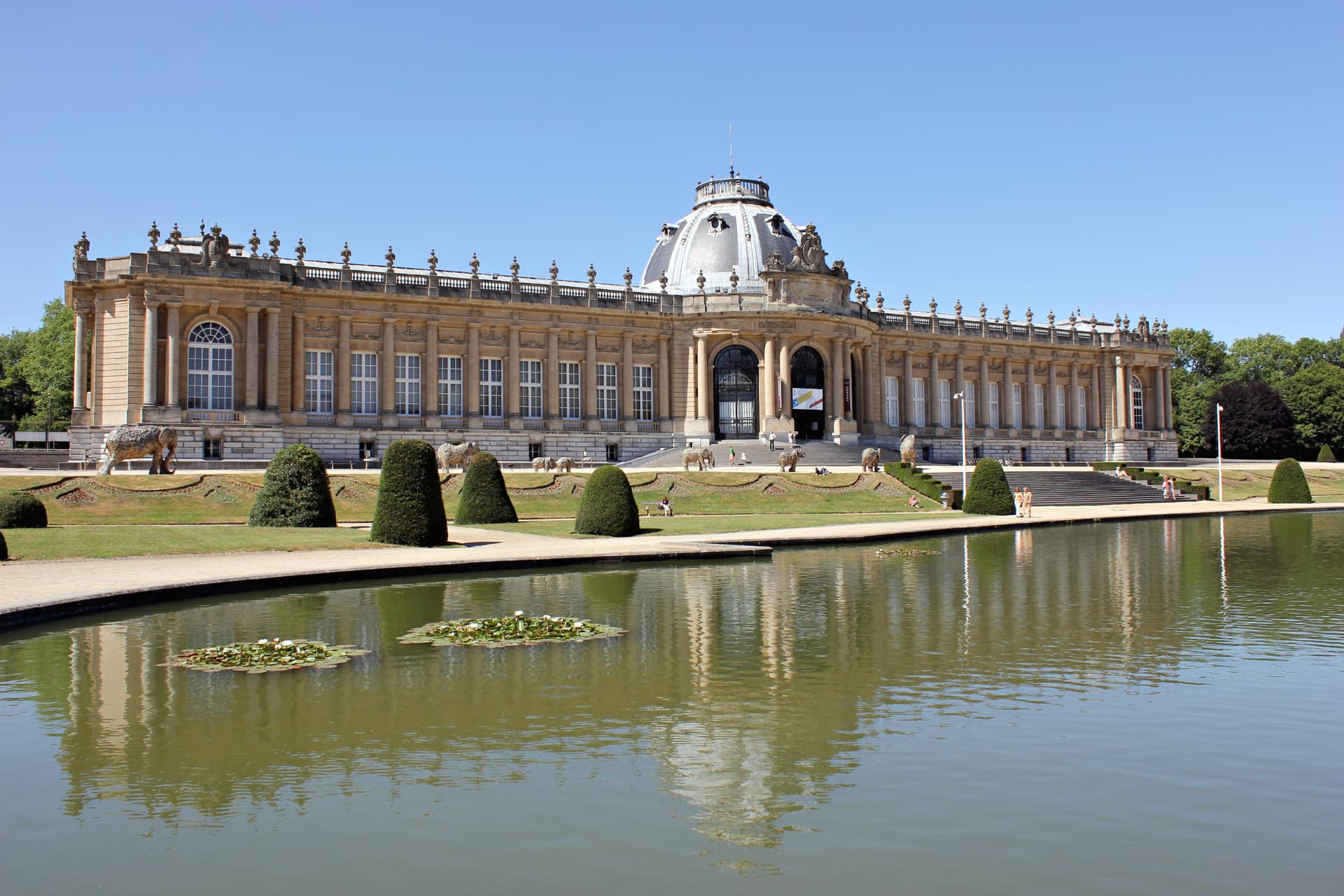 Royal Museum for Central Africa building reflected in pond with water lilies and elephant sculptures.