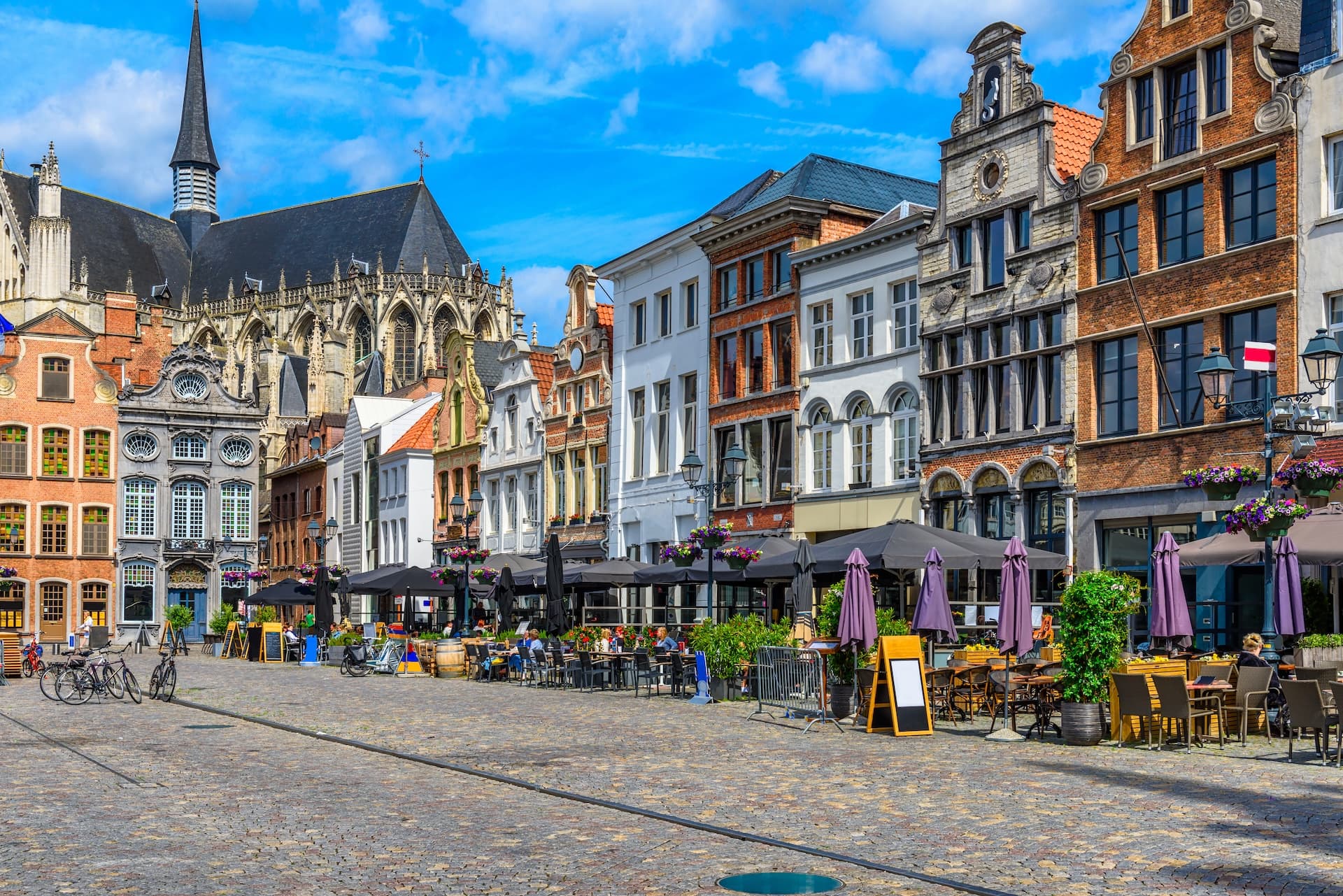 Cobblestone square with outdoor cafe seating and historic buildings, including a church spire in Mechelen.