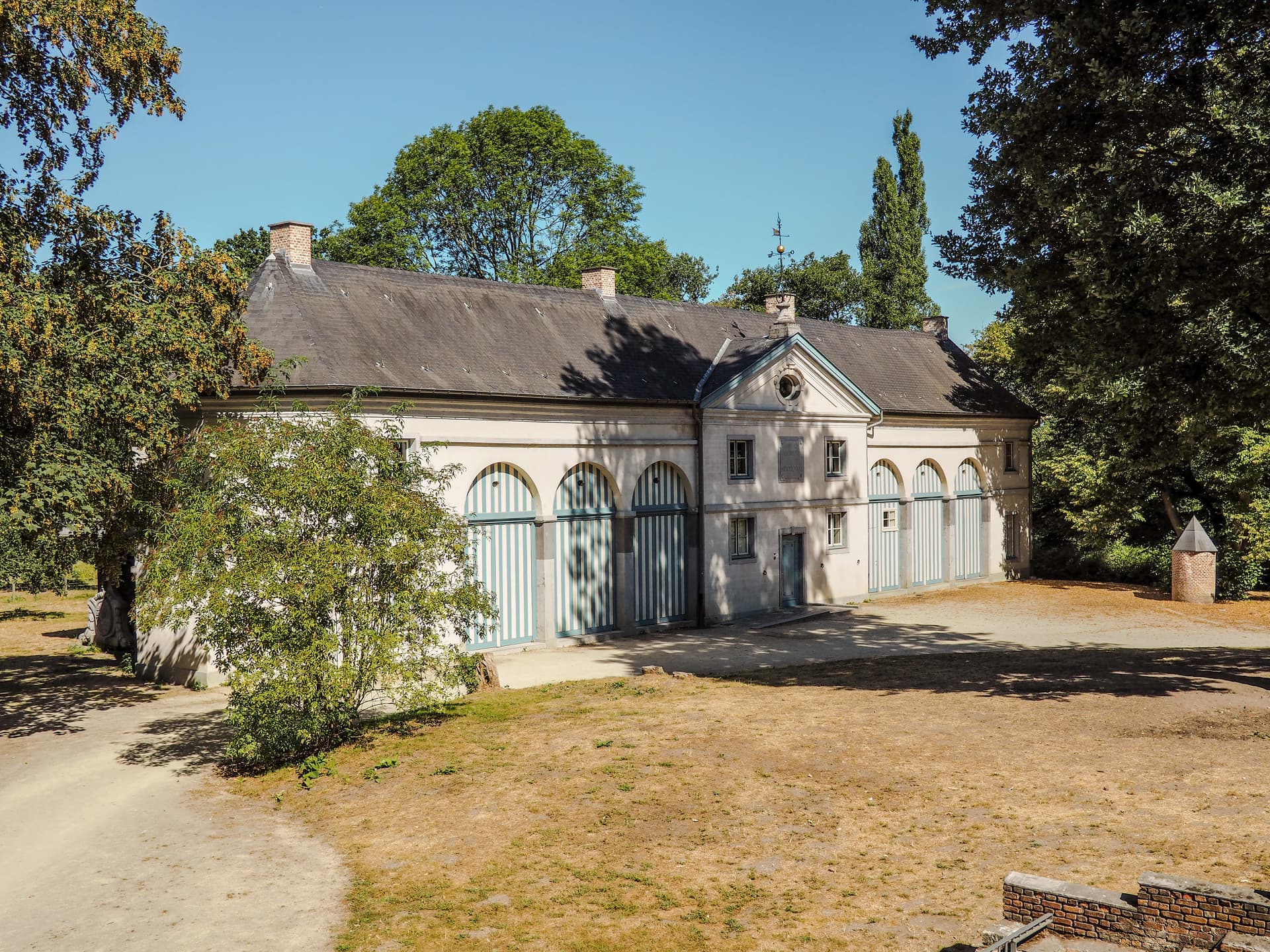 Historic building with striped arched doors, weathervane, and dry lawn under clear blue sky.