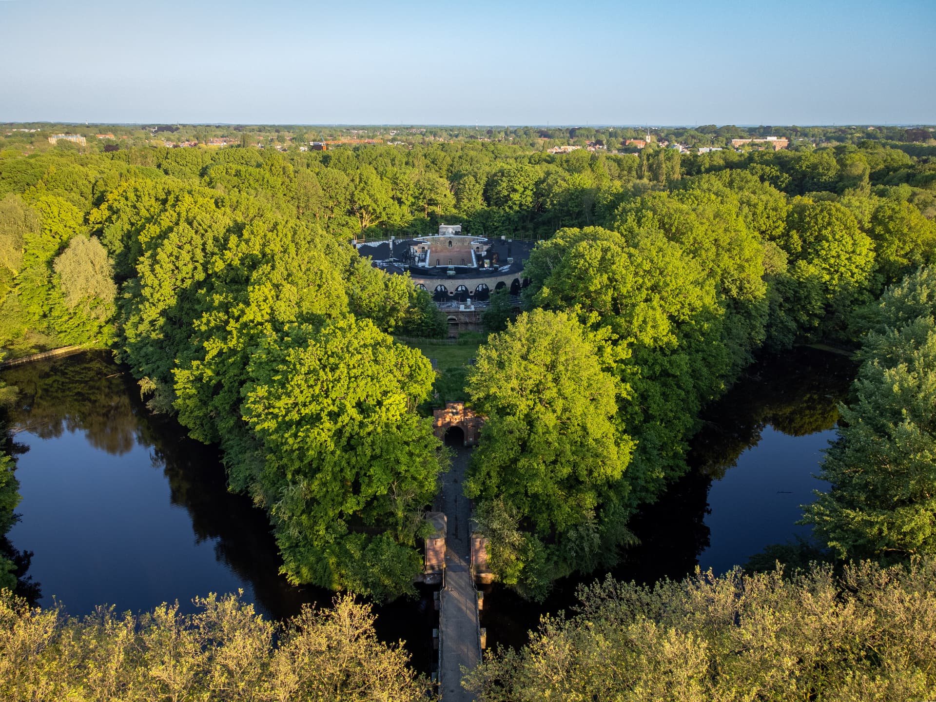 Fortress structure surrounded by dense green trees and dark water moat under a clear blue sky.