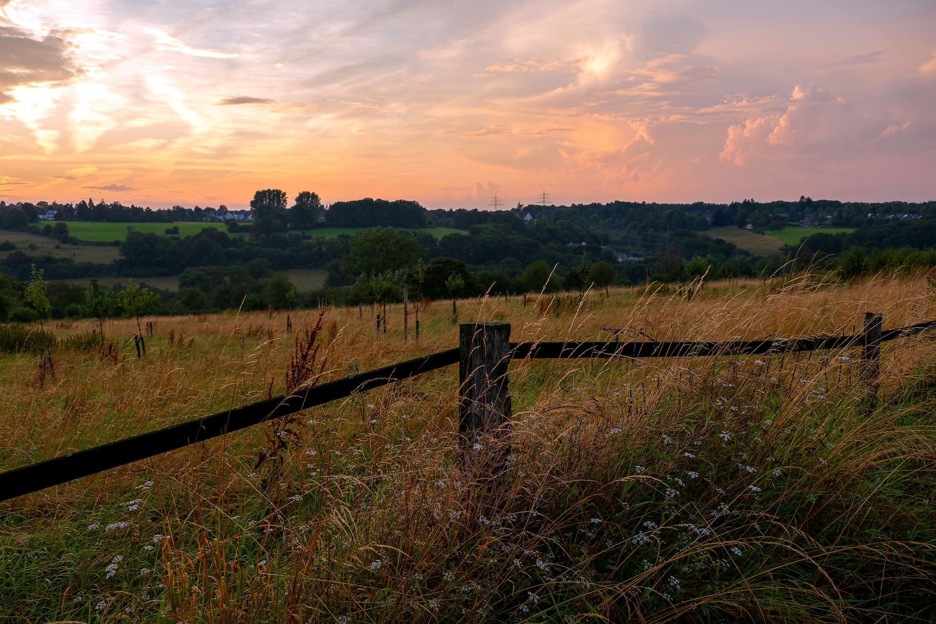 Grassy field with wooden fence in foreground and rolling hills at sunset
