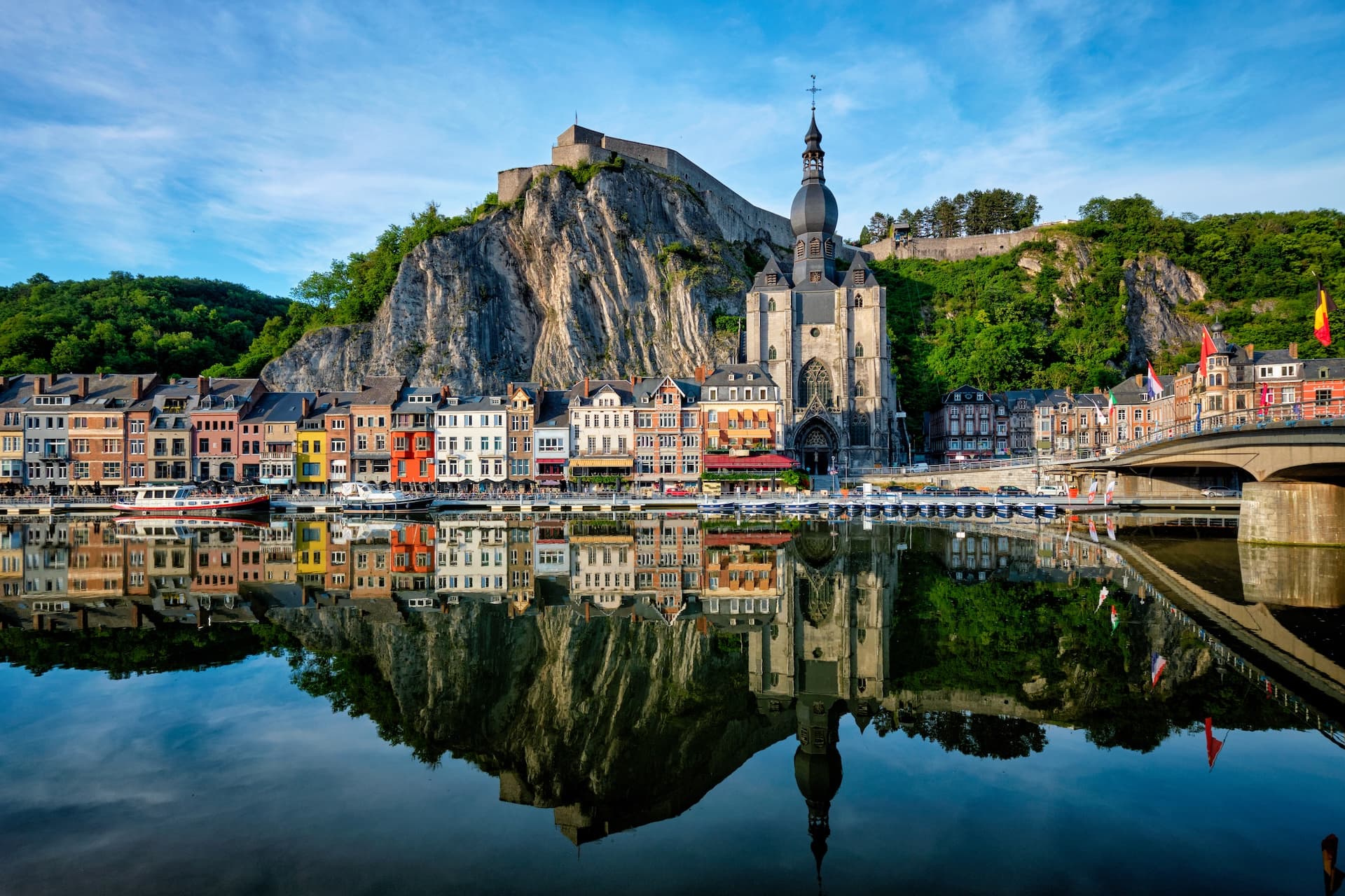 Colorful town reflected in calm river below steep cliff with citadel and church in Dinant, Belgium.