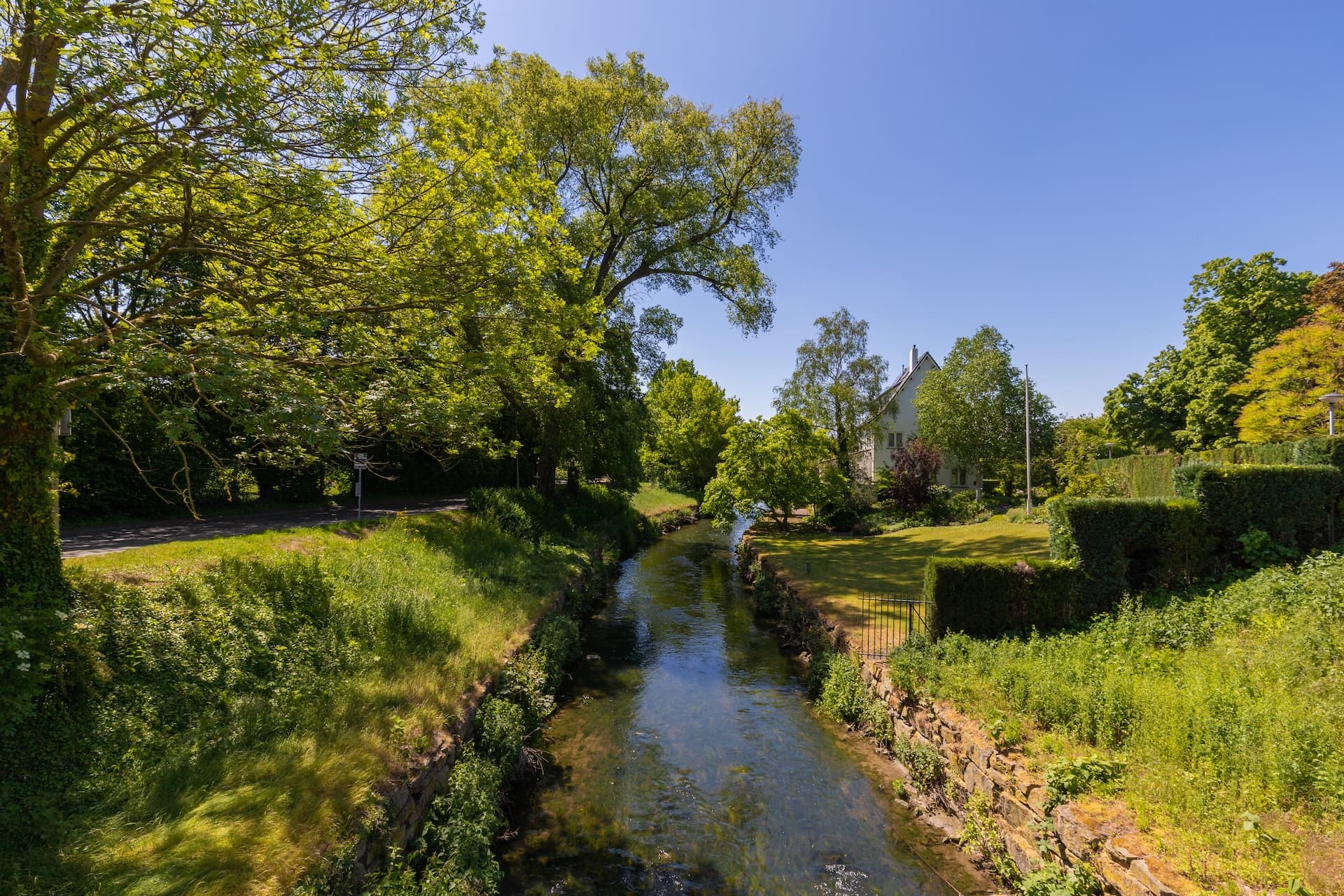 Clear stream flowing through lush green banks past a white house under a bright blue sky.