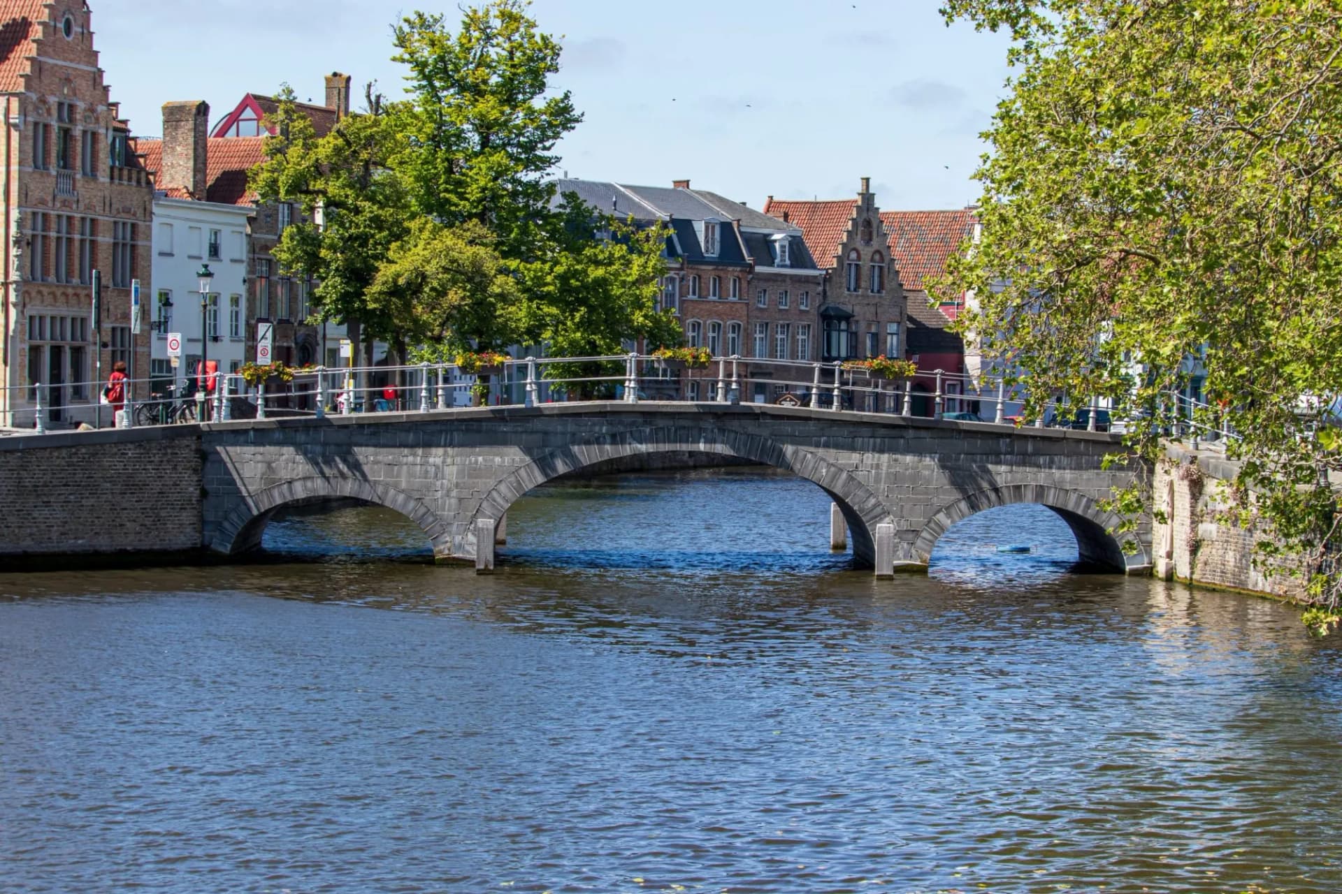 Stone arch bridge over canal with historic buildings and green trees in Bruges, Belgium.