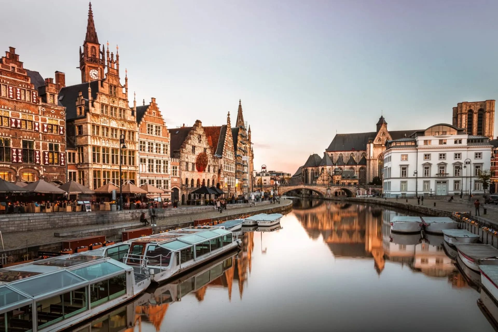 Historic buildings line the canal with tour boats docked in Ghent, Belgium at dusk.