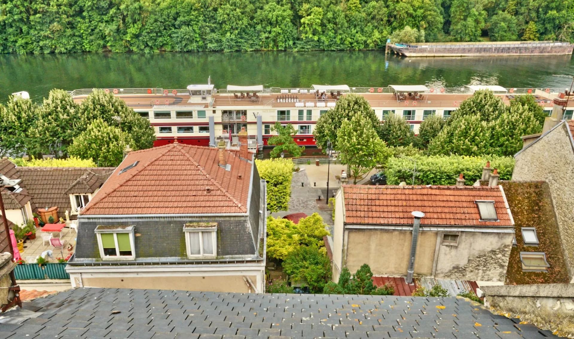 Rooftops overlooking a river with a passenger boat and barge in Conflans-Sainte-Honorine, France.