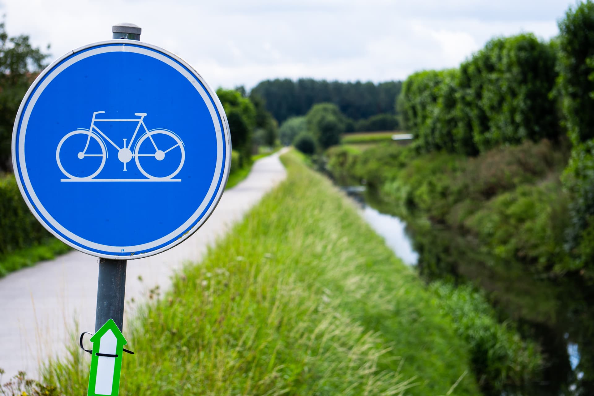Mandatory bicycle path sign next to a paved trail alongside a narrow canal with lush greenery.